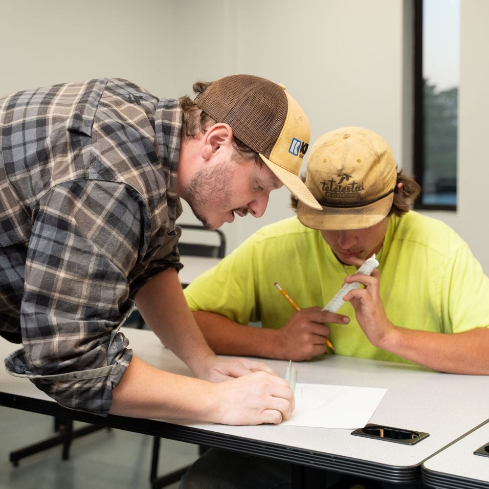 Two men sitting at a desk in a classroom work together on a paper. One, wearing a plaid shirt and cap, leans over to explain something to the other, who is wearing a yellow shirt and holding a pencil and ruler.