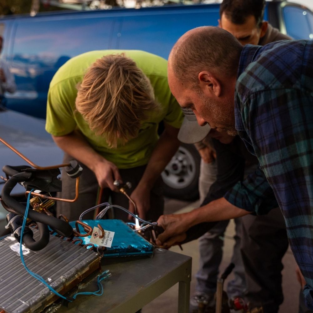 Two people closely examine and work on an open electronic device with wires and components on a metal table outdoors, with a blue van parked in the background.