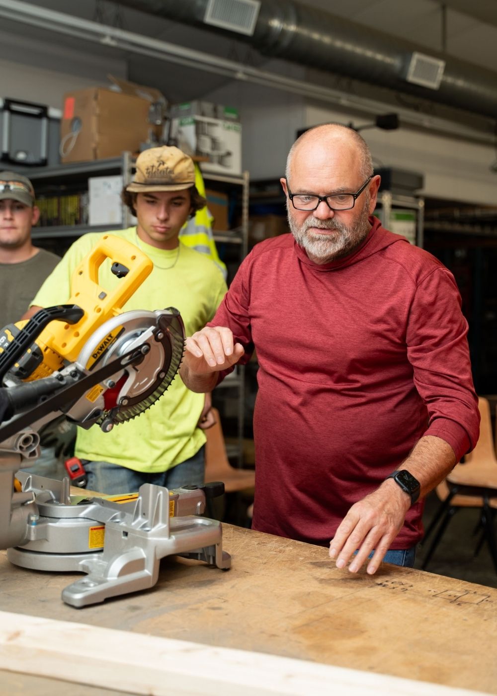 A man in a red shirt stands next to a miter saw on a workbench, with two younger men in work clothes and safety hats watching in the background in a workshop.