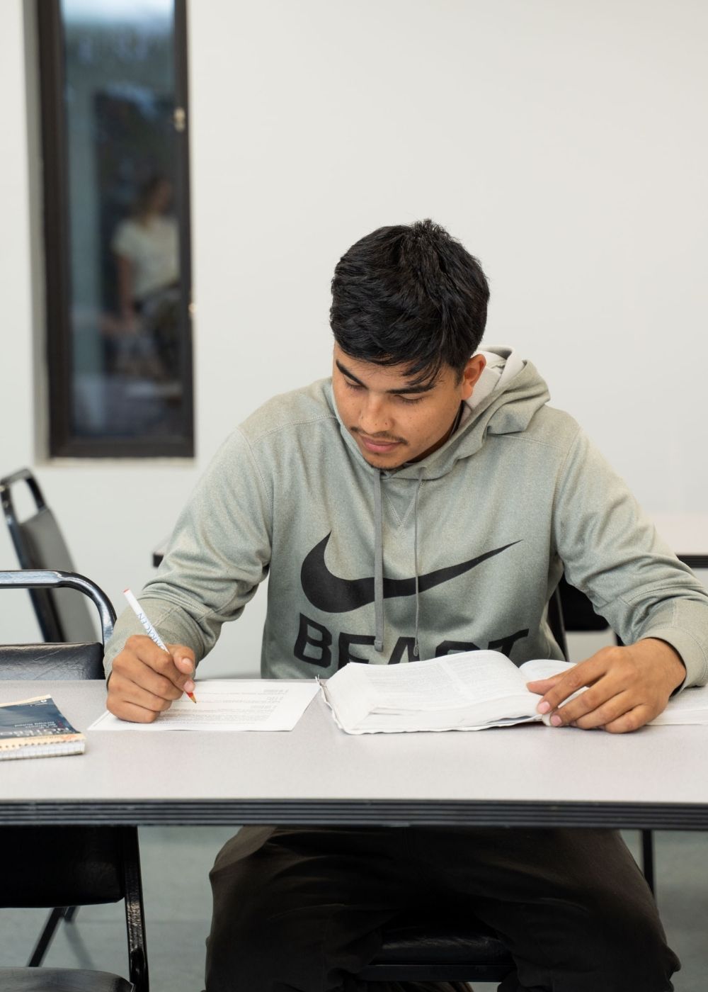 A young man wearing a gray hoodie is sitting at a desk in a classroom, writing on paper with a pen while looking at an open notebook. There are extra chairs and a window in the background.