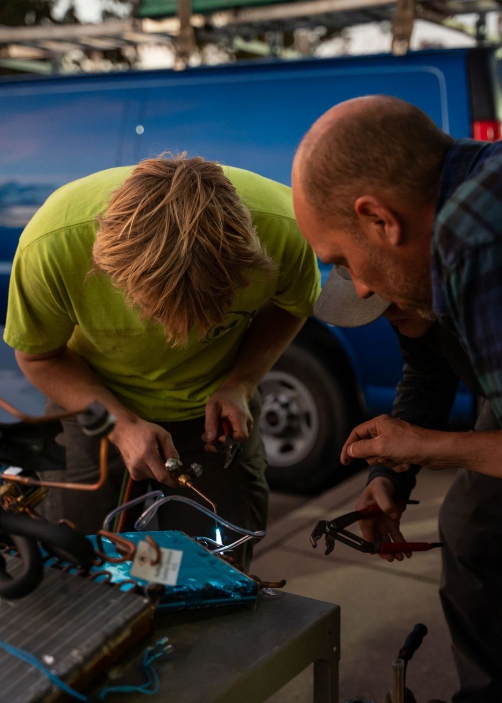 Two people bend over a metal workbench, using tools to work on a mechanical project outdoors. A blue van is parked in the background. The focus is on the activity and teamwork.