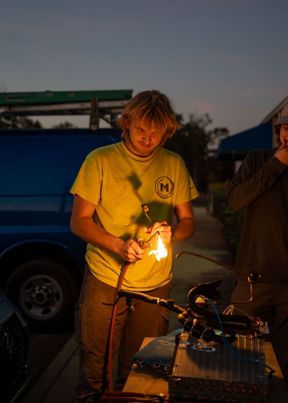 A person with blonde hair in a yellow shirt is working outdoors at dusk, holding a lit blowtorch over equipment on a table. A blue van and another person are visible in the background.