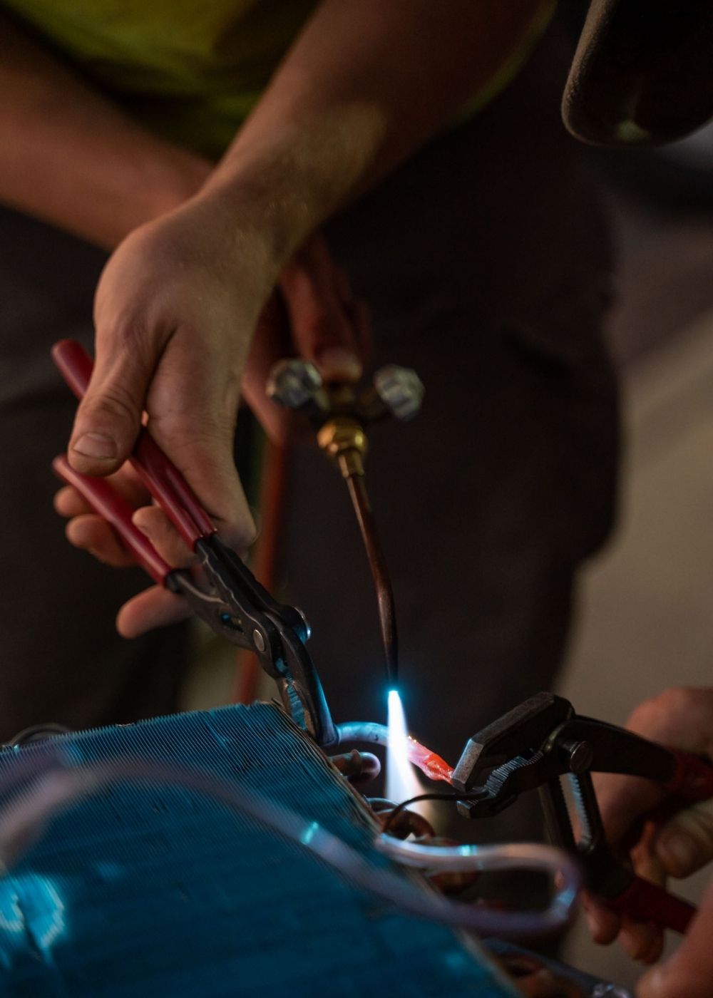A close-up of hands using pliers and a blowtorch to work on metal components, possibly during a repair or welding process. Blue flame and tools are clearly visible, with focused attention on the task.