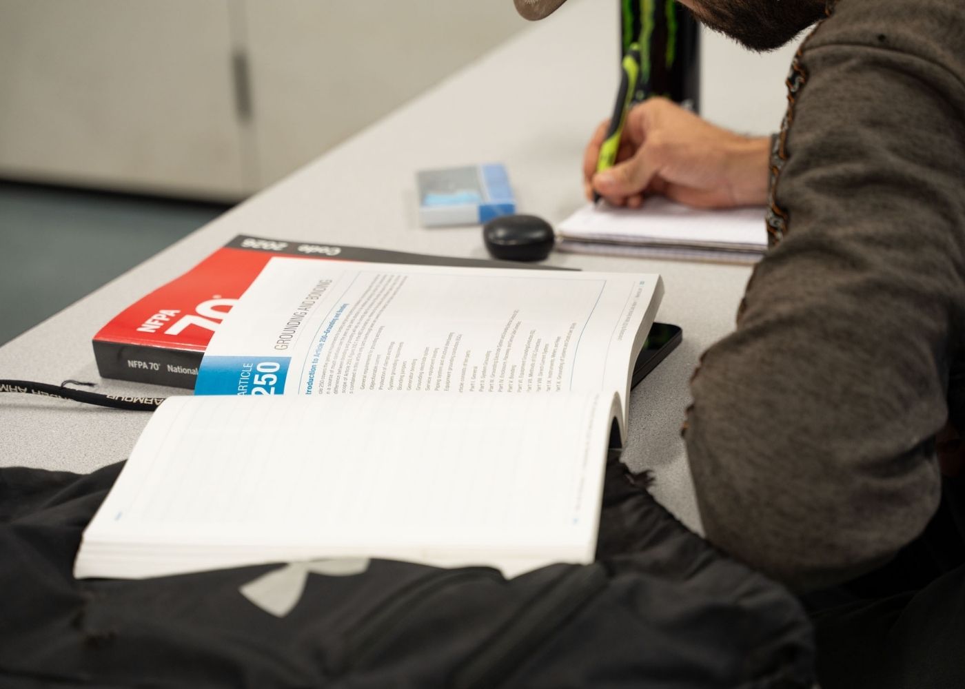 A person sits at a desk, highlighting notes in a notebook. An open textbook and a closed PSAT/NMSQT prep book lie on the table, along with a black backpack and a pen.