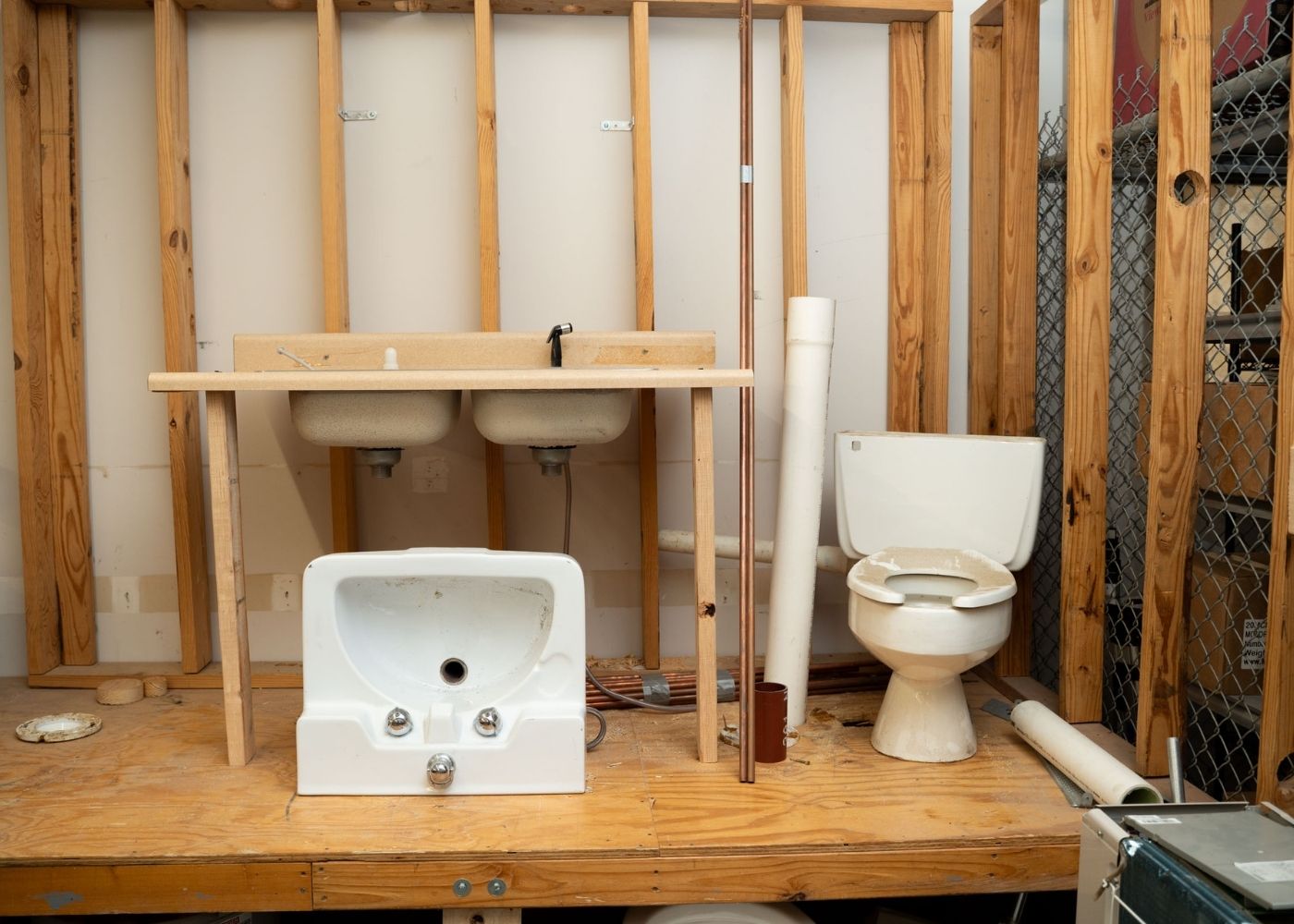 A partially built bathroom setup with exposed wooden wall studs, a double sink, a detached wall-mounted sink, a toilet, and various pipes placed on a wooden platform.