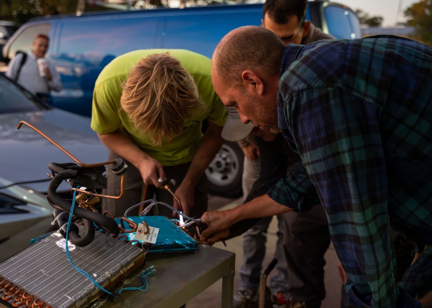 Several people work together on an outdoor project, assembling or repairing an electronic device with wires and metal components on a table, with a blue van and car in the background.