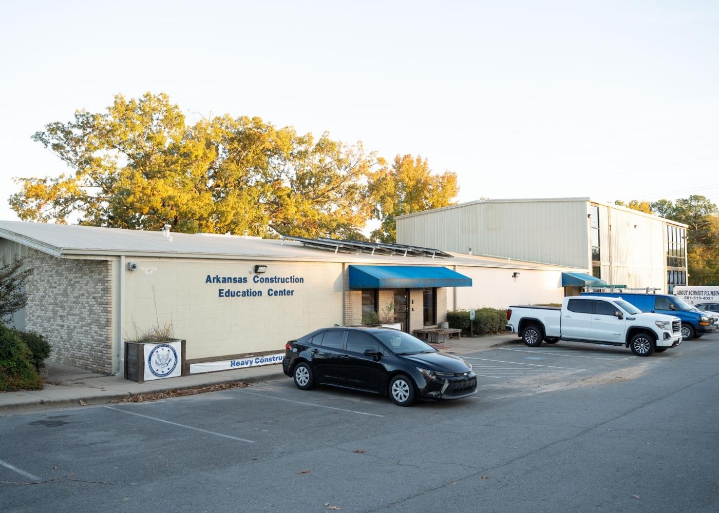 A single-story building with a sign reading Arkansas Construction Education Center and several vehicles parked in front, surrounded by trees with autumn foliage.