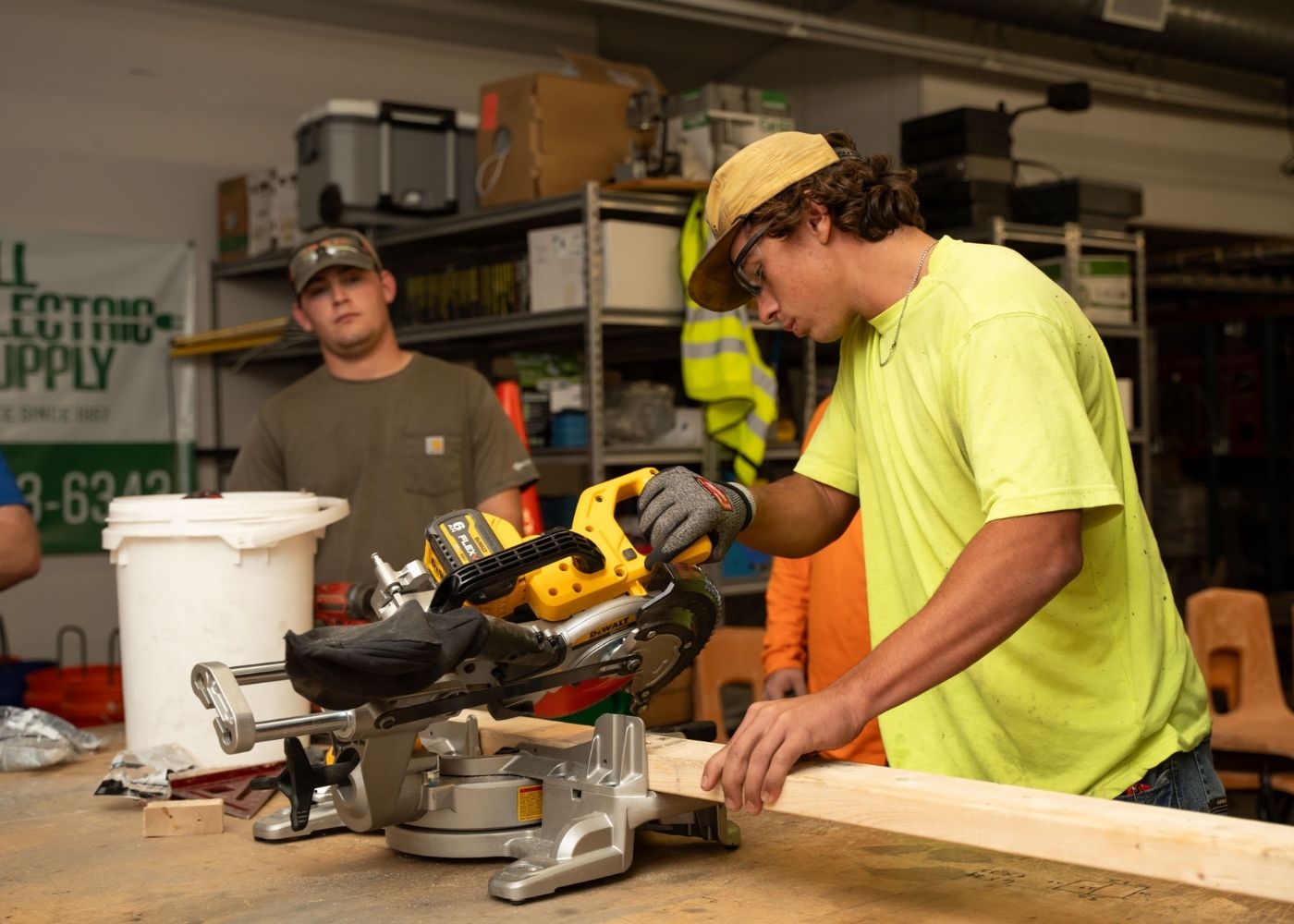 A man in a yellow shirt and cap uses a miter saw to cut a wooden plank in a workshop, while another man in a brown shirt watches. Shelves with tools and supplies are visible in the background.