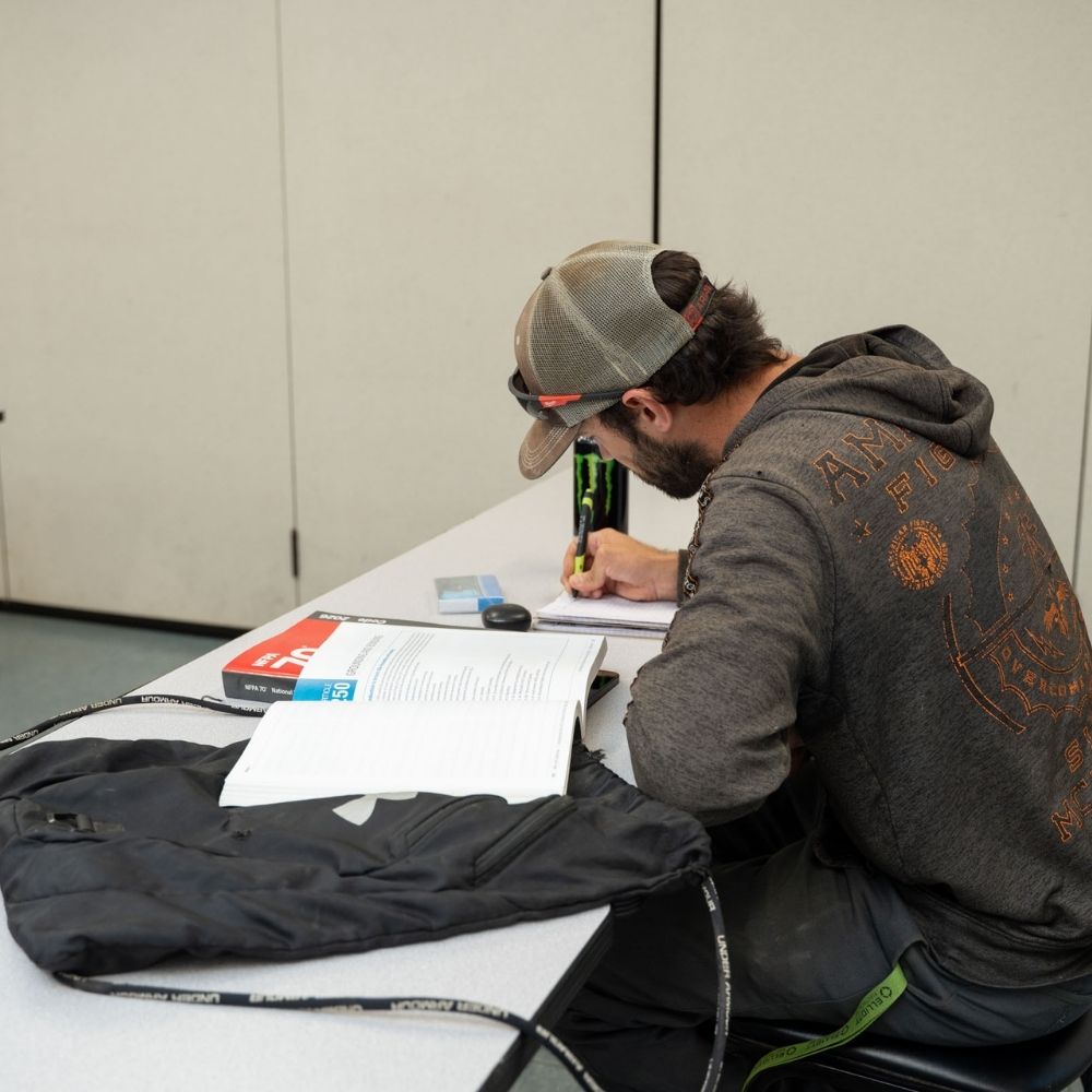 A person wearing a cap and hoodie sits at a table, writing in a notebook with a can of Monster energy drink nearby. An open textbook, pen, and backpack are also on the table.