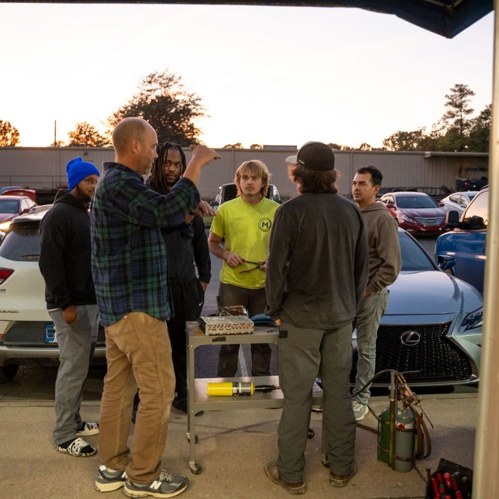 A group of six men stand in a parking lot, gathered around a small table with tools and gas tanks. Cars are parked behind them, and the sun is setting in the background. One man gestures as he speaks to the group.
