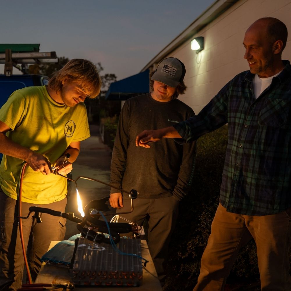 Three people stand outside near a building at dusk. One person uses a torch, creating a bright flame, while the others watch and smile. Equipment and tools are set up on a table in front of them.