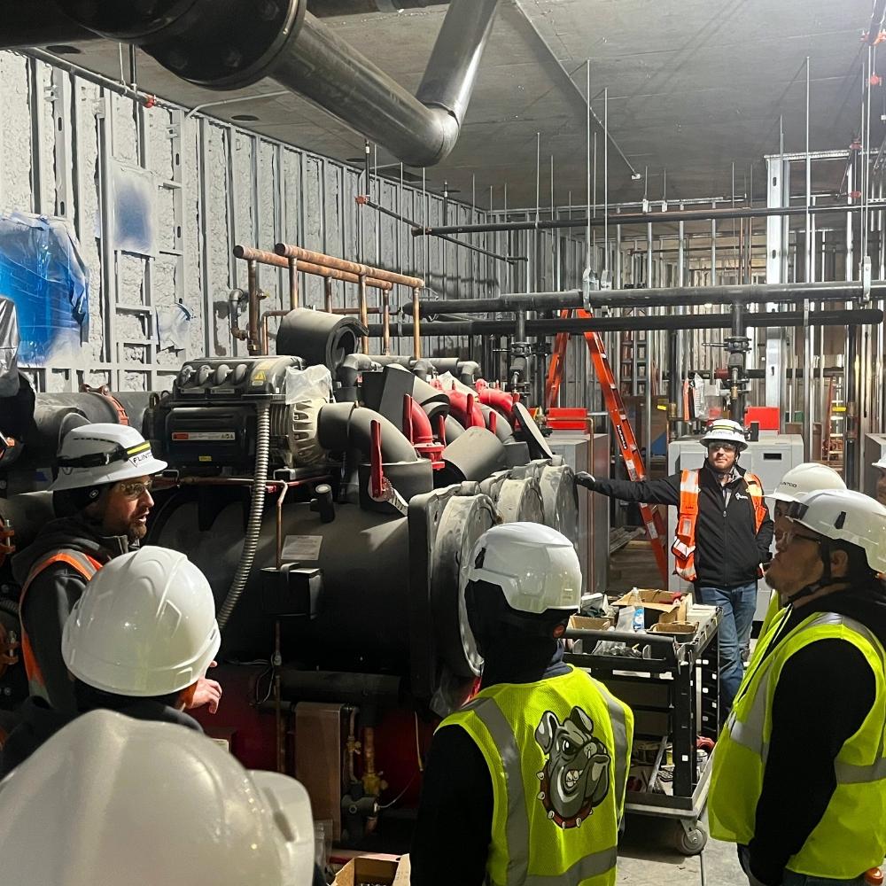 Several construction workers in safety vests and hard hats gather around industrial equipment inside a partially finished building, discussing the machinery. Exposed pipes, wiring, and construction materials are visible.