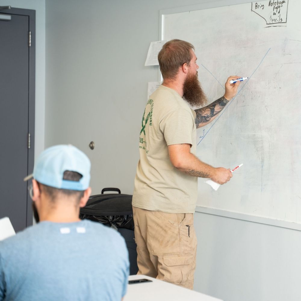 A man with a beard writes on a whiteboard with blue marker in a classroom, while another person in a blue shirt and cap sits facing the board, taking notes.