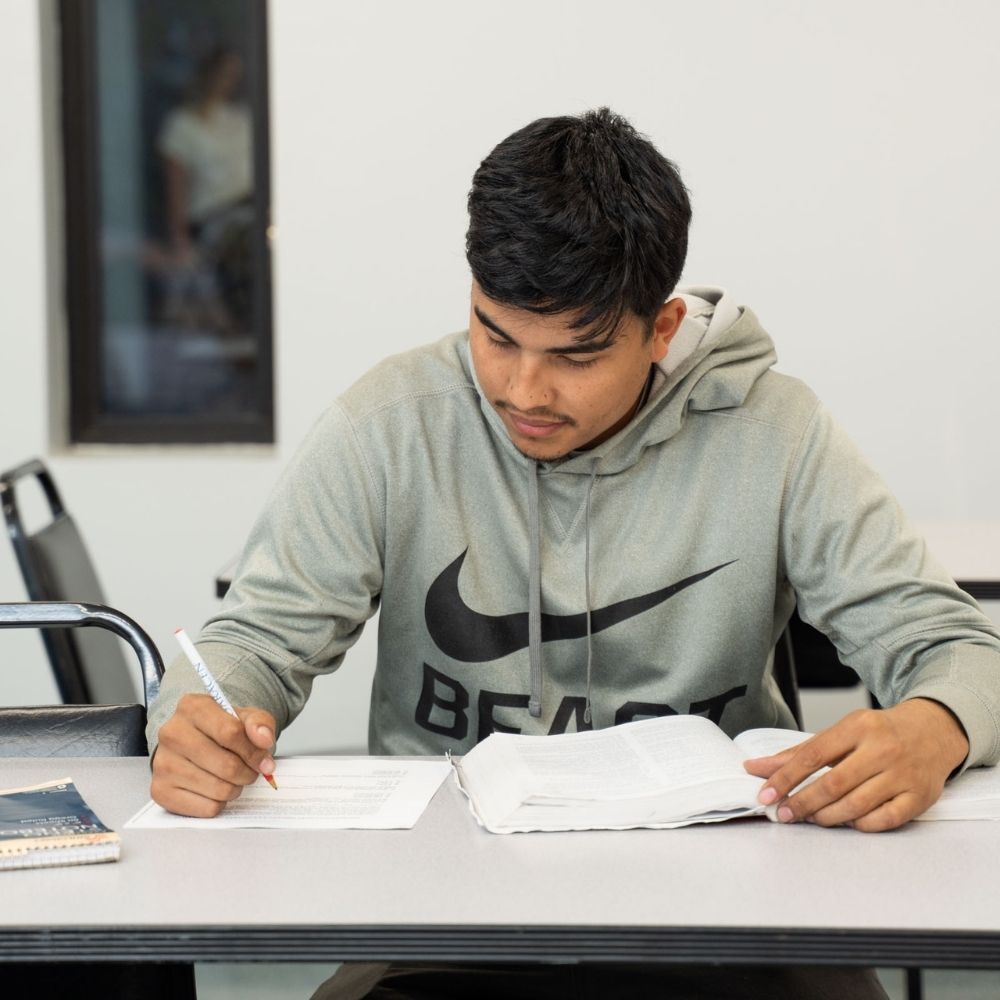 A young man in a gray Nike hoodie is sitting at a desk, writing on a sheet of paper with a pen while reading an open book. A notebook is also on the desk, and the background is a plain classroom setting.