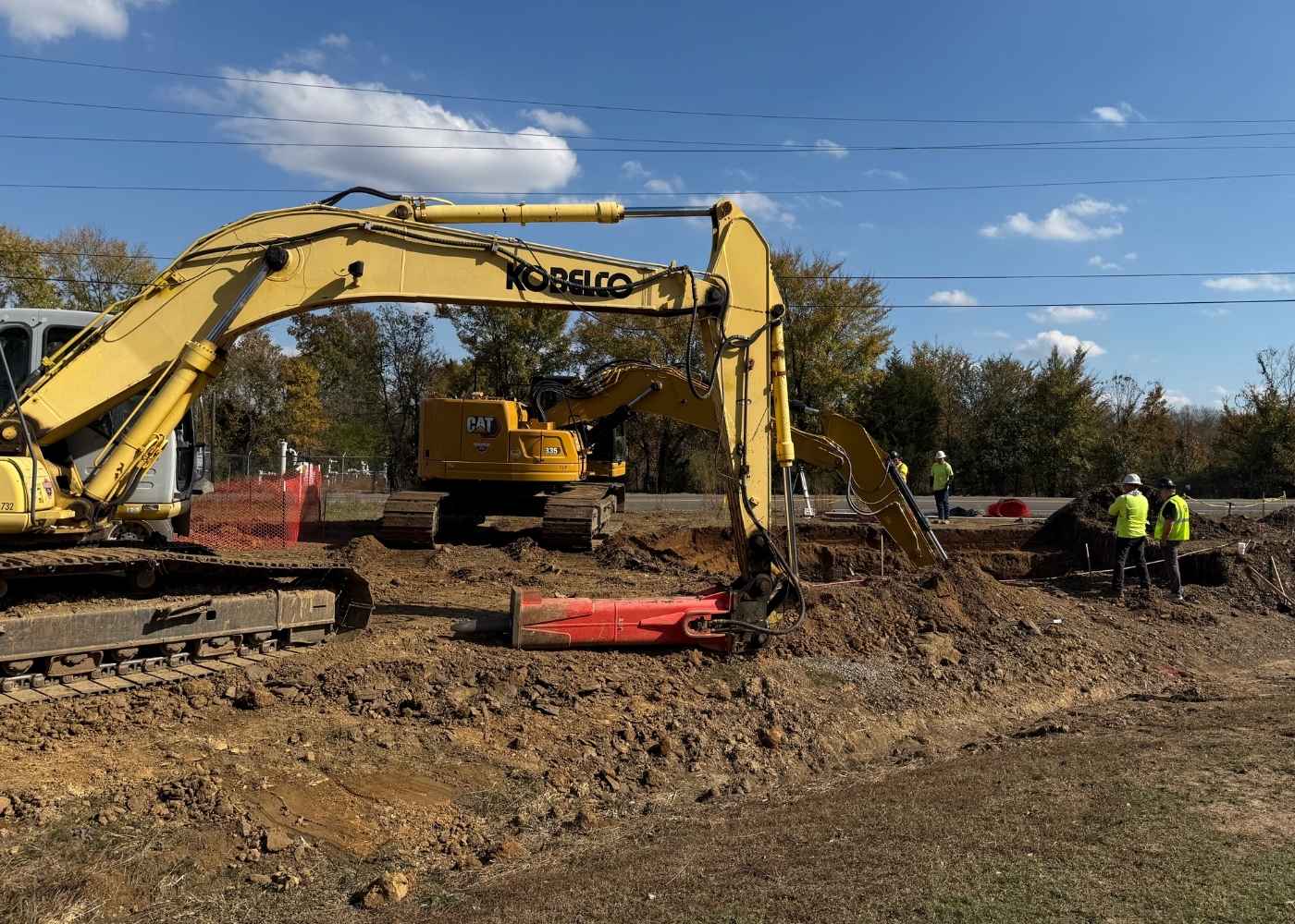 Two large yellow excavators work at a construction site while several workers in safety vests and helmets stand nearby. The ground is dirt and rubble, with trees and power lines in the background under a blue sky.