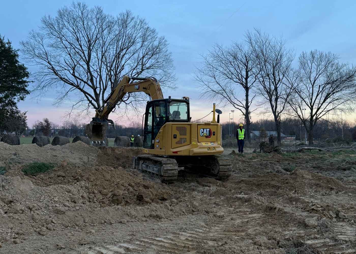 A yellow excavator digs soil at a construction site as two workers in safety vests observe nearby. Leafless trees and a cloudy sky are visible in the background.