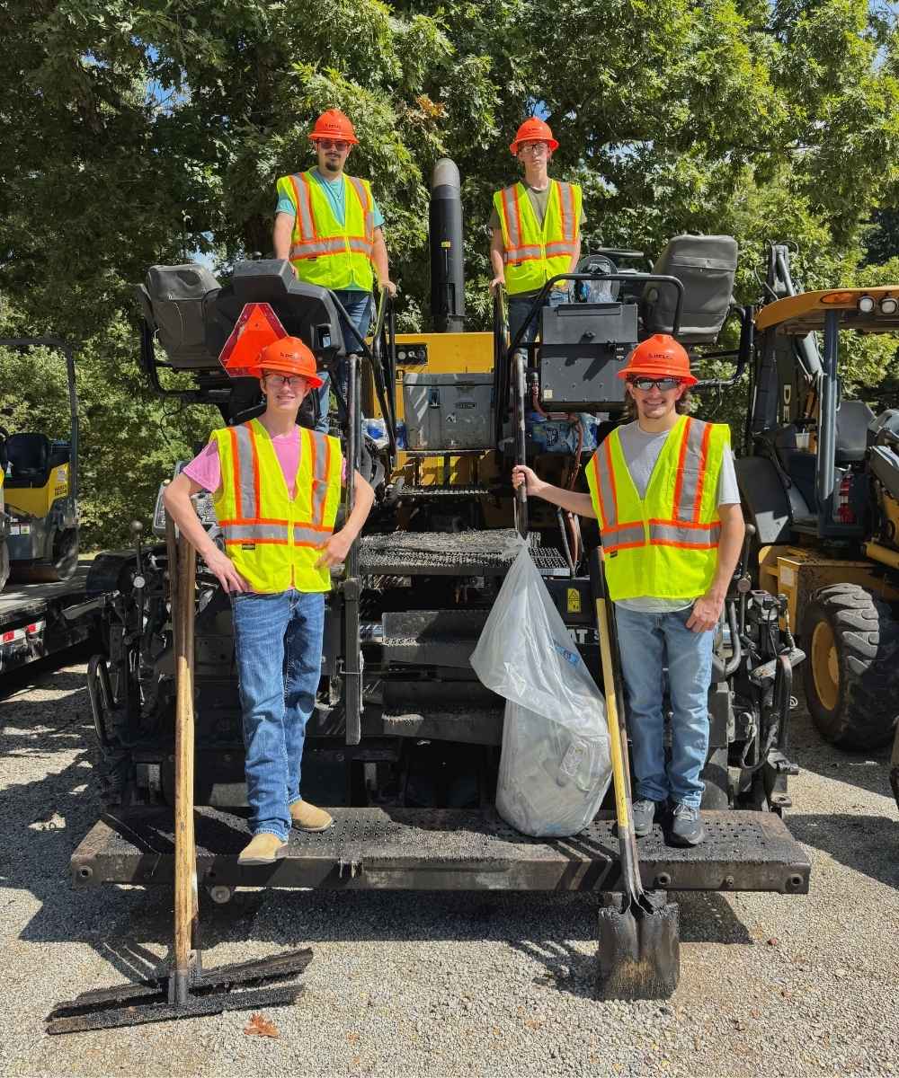 Four construction workers in yellow safety vests and orange hard hats stand on and in front of heavy machinery outdoors, holding shovels and a trash bag, with trees and more equipment in the background.