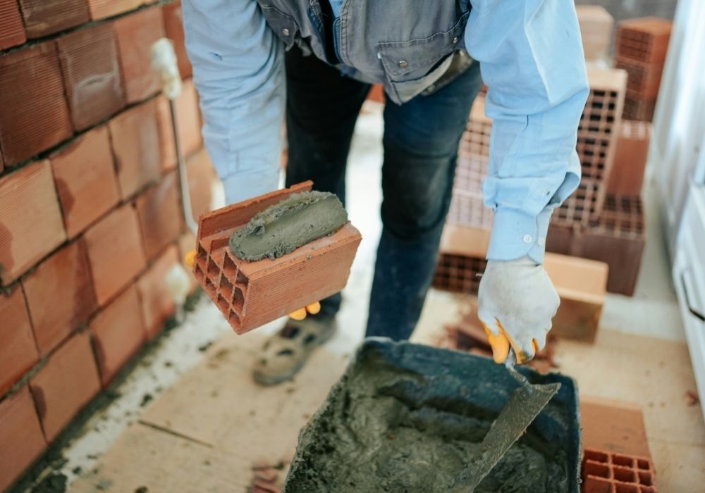 A construction worker wearing gloves spreads mortar on a red brick with a trowel, preparing to lay it on a partially built brick wall in an indoor setting. More bricks and a container of mortar are visible nearby.