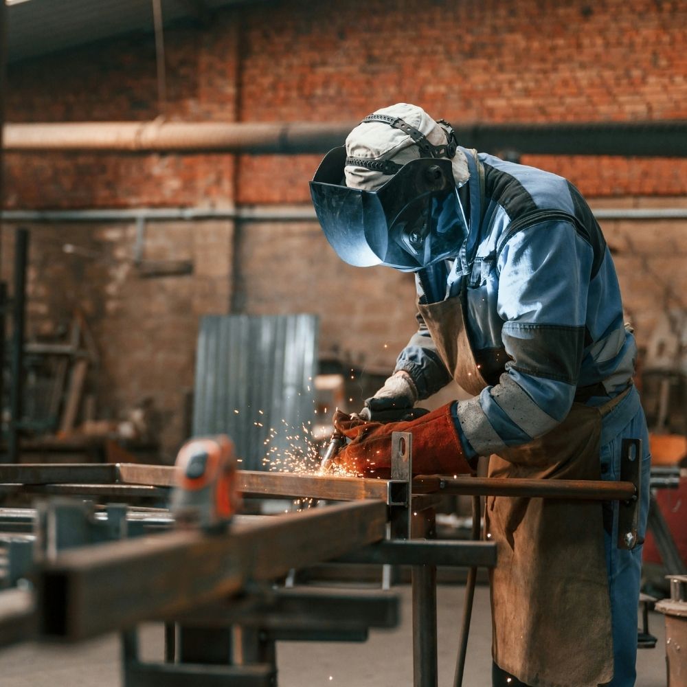 A worker in protective gear, including a welding mask and gloves, welds metal in an industrial workshop, with sparks flying from the welding process. The background shows tools and a brick wall.