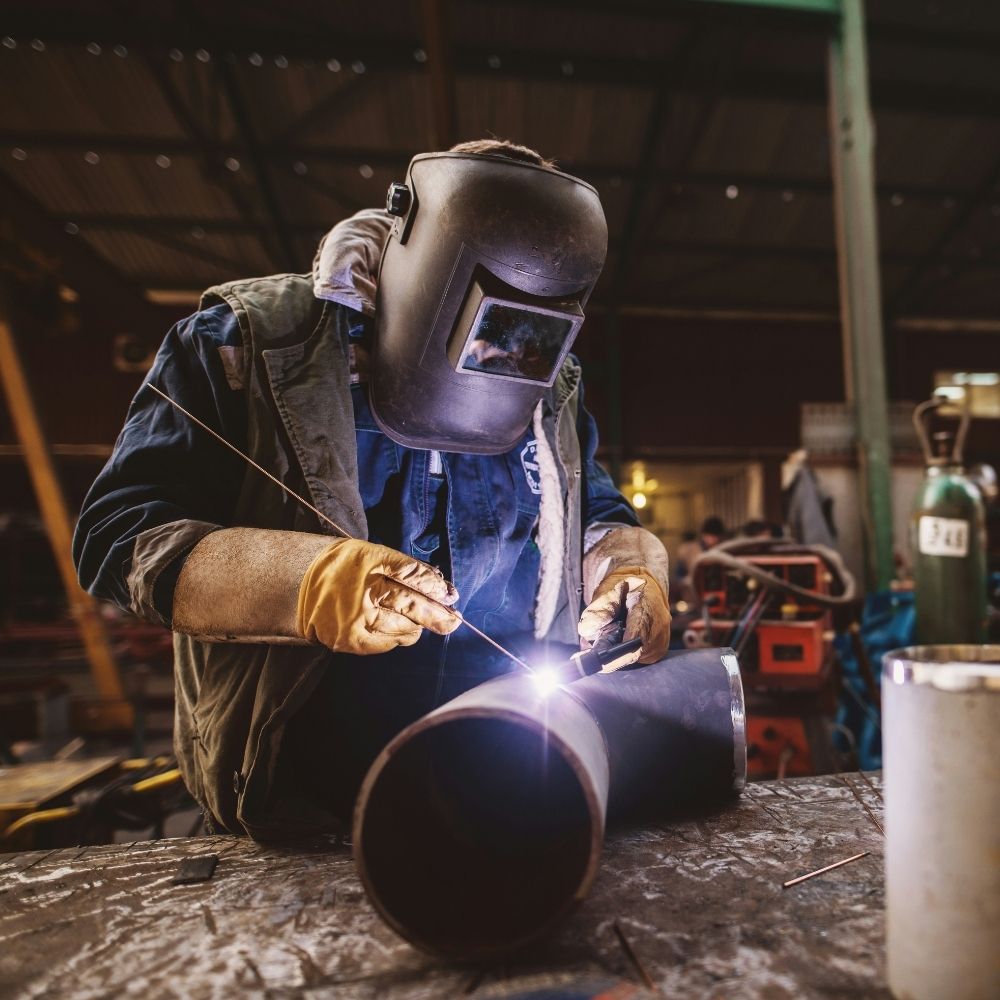 A person wearing protective gloves and a welding helmet uses a welding torch to join metal pipes on a workbench in an industrial workshop.