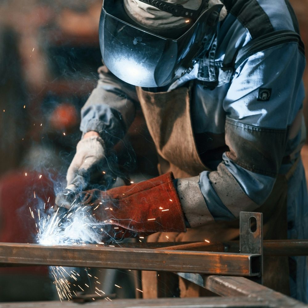 A person wearing protective gear, including a welding mask and gloves, welds metal beams, producing bright sparks and smoke in an industrial setting.