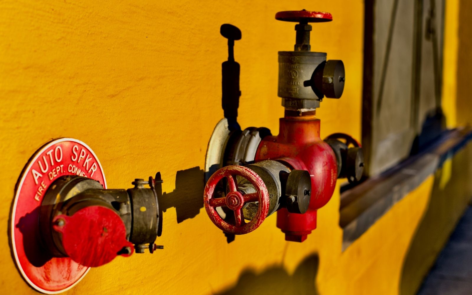 A close-up of a red fire department connection valve and hydrant mounted on a bright yellow wall, with a shadow cast to the right and a window partially visible in the background.