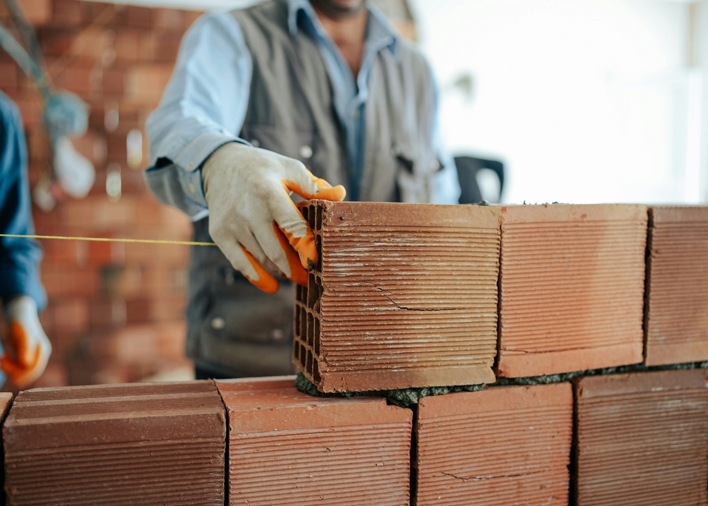 A construction worker wearing gloves carefully places a red brick on a wall, aligning it with a yellow string for accuracy. Other bricks and blurred workers are visible in the background.
