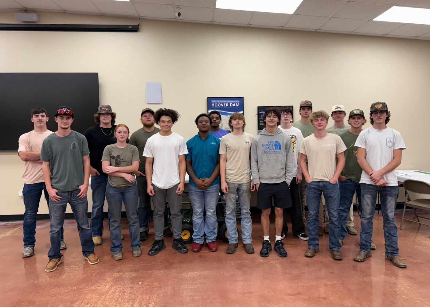 A group of sixteen young people, mostly male, stand indoors in a classroom with beige walls, some smiling at the camera. They wear casual clothes including jeans, T-shirts, and work boots. A Hoover Dam poster is behind them.
