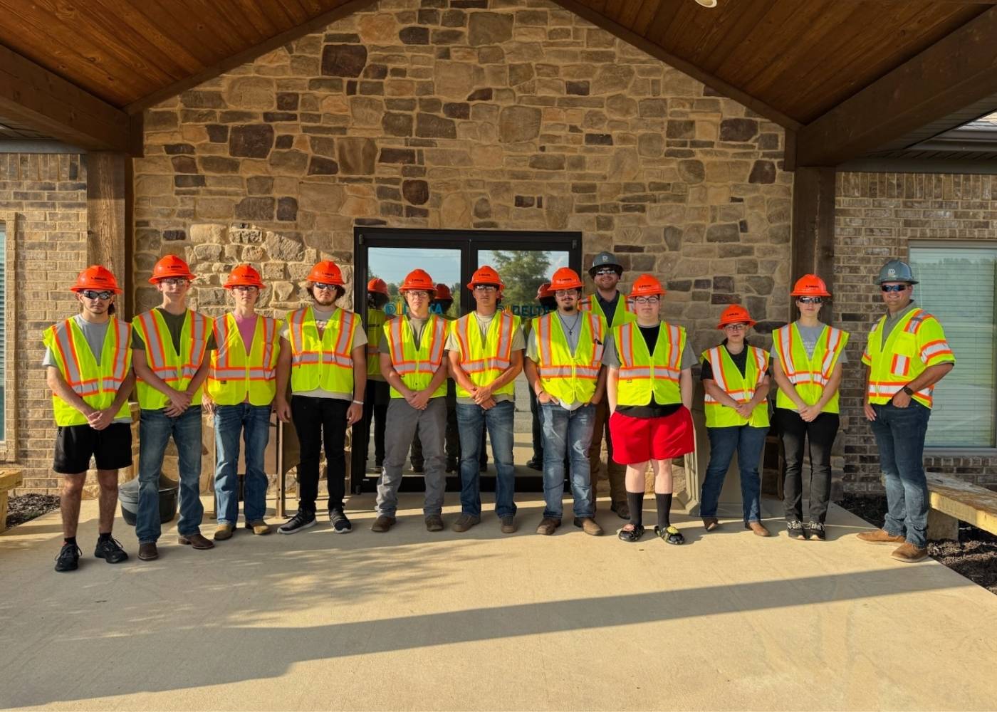 A group of thirteen people wearing orange hard hats and yellow safety vests stand in a line outside a stone building, posing for a group photo in front of a glass door.