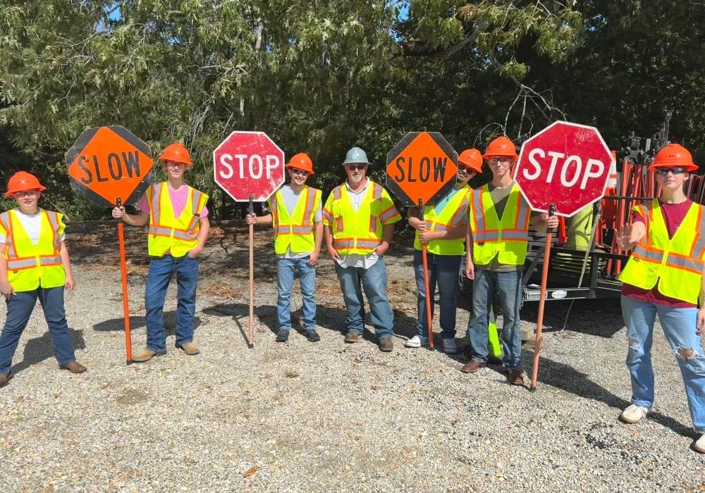 A group of seven people wearing safety vests, hard hats, and sunglasses stand outdoors on gravel holding “STOP” and “SLOW” signs, with trees visible in the background.
