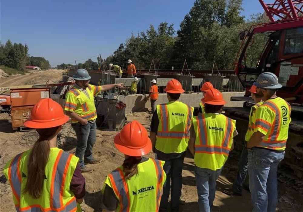 A group of construction workers in yellow safety vests and orange helmets listen to a supervisor pointing at equipment on a construction site with machinery and concrete barriers in the background.