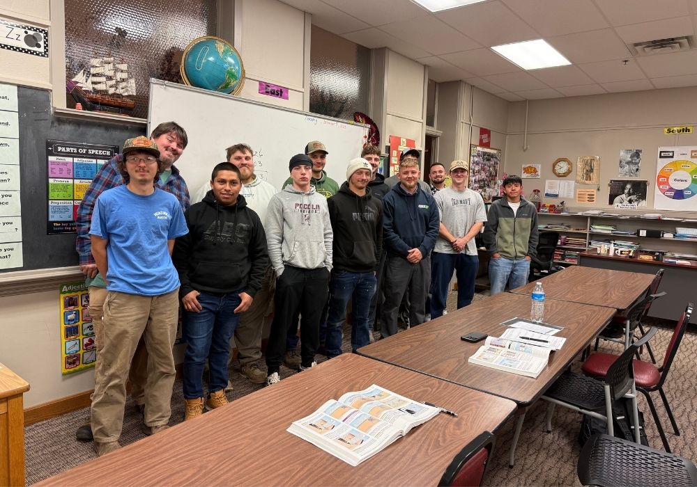 A group of twelve people stand side by side in a classroom next to long tables with open books, posters, and educational materials on the walls and shelves in the background.