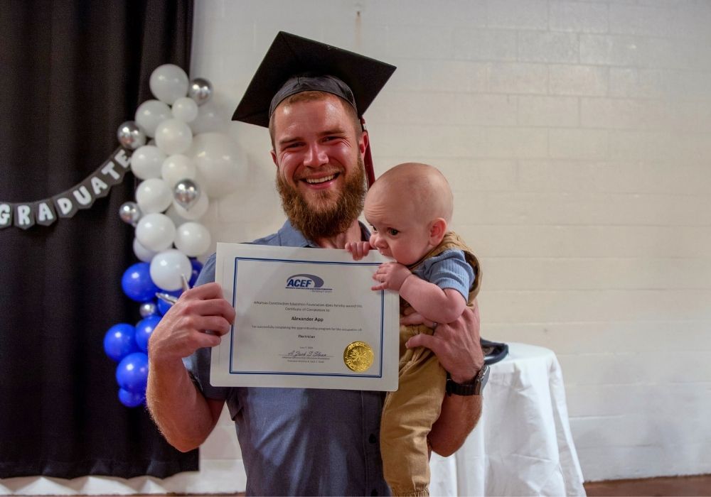 A smiling man in a graduation cap holds a certificate in one hand and a baby in the other. Behind him are white and blue balloon decorations and a black banner with GRADUATE spelled out.