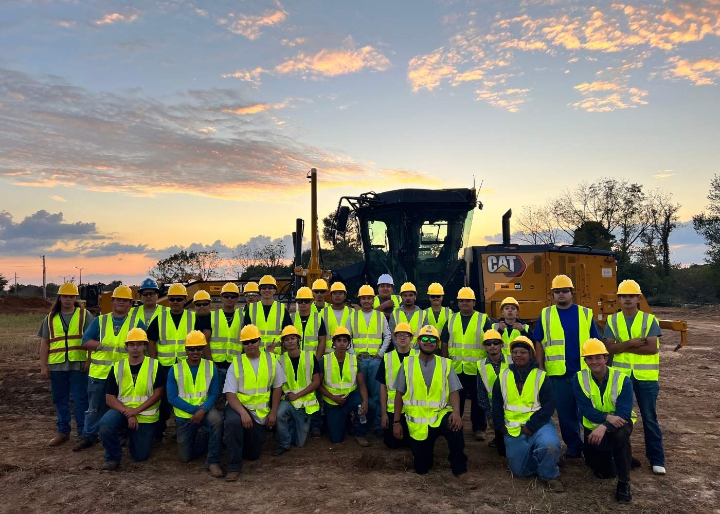 A group of construction workers wearing yellow safety vests and hard hats pose for a group photo in front of a large construction vehicle at a worksite during sunset.