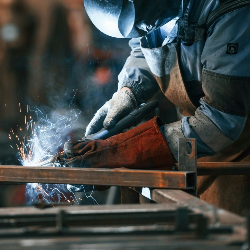 A person wearing protective gloves and a welding helmet uses a welding torch on a metal frame, with sparks and smoke visible in the workshop environment.