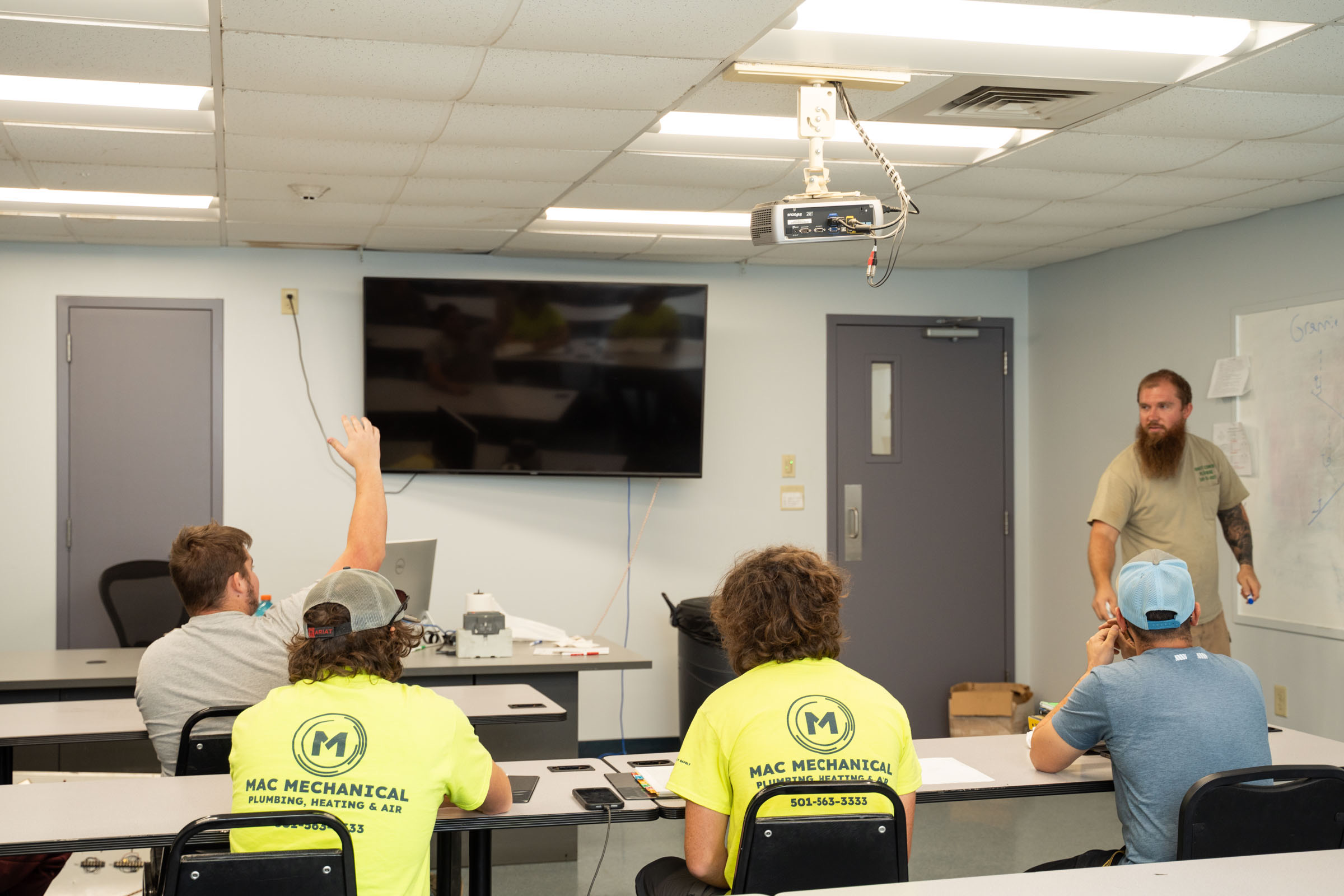 A bearded man stands at the front of a classroom speaking, while four people wearing “MAC MECHANICAL” shirts sit at desks. One person raises their hand. A large TV and a projector are mounted on the ceiling.