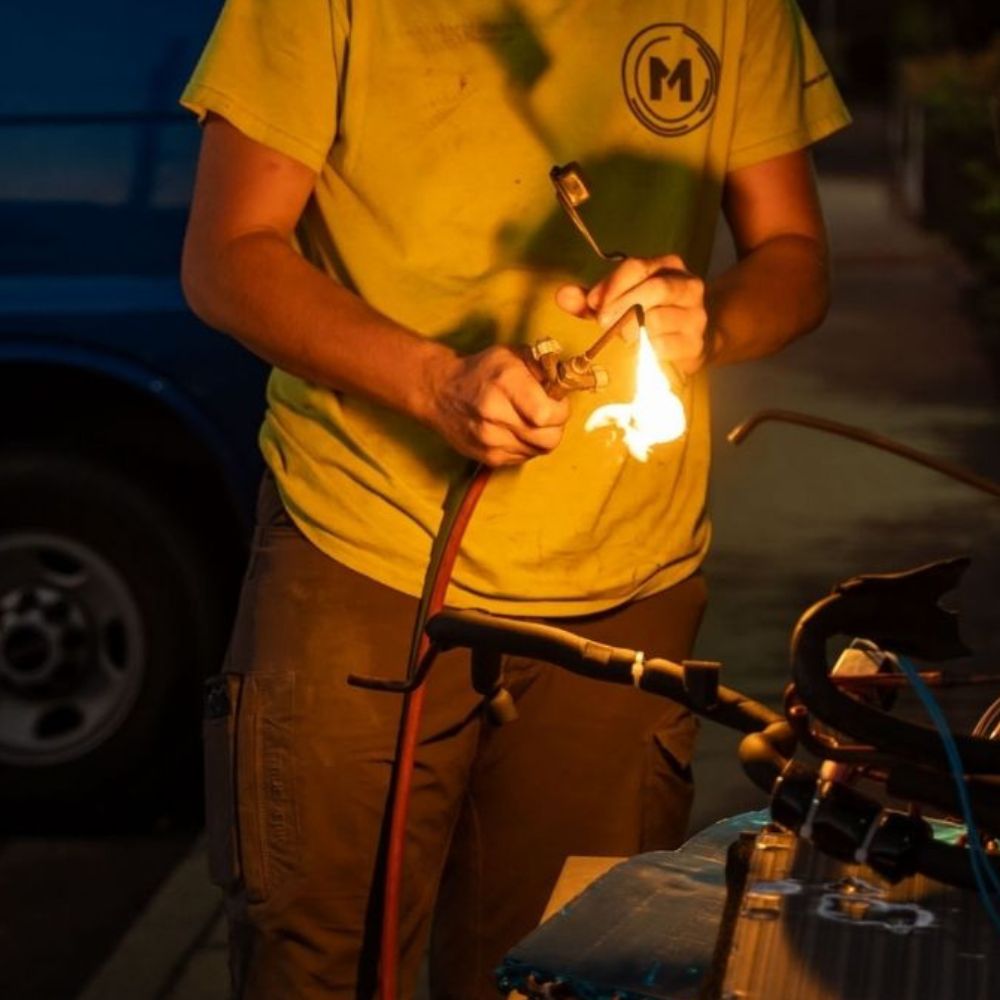 Person in a yellow shirt using a torch to work with fire on metal equipment outside at night, with tools and a blue van in the background.